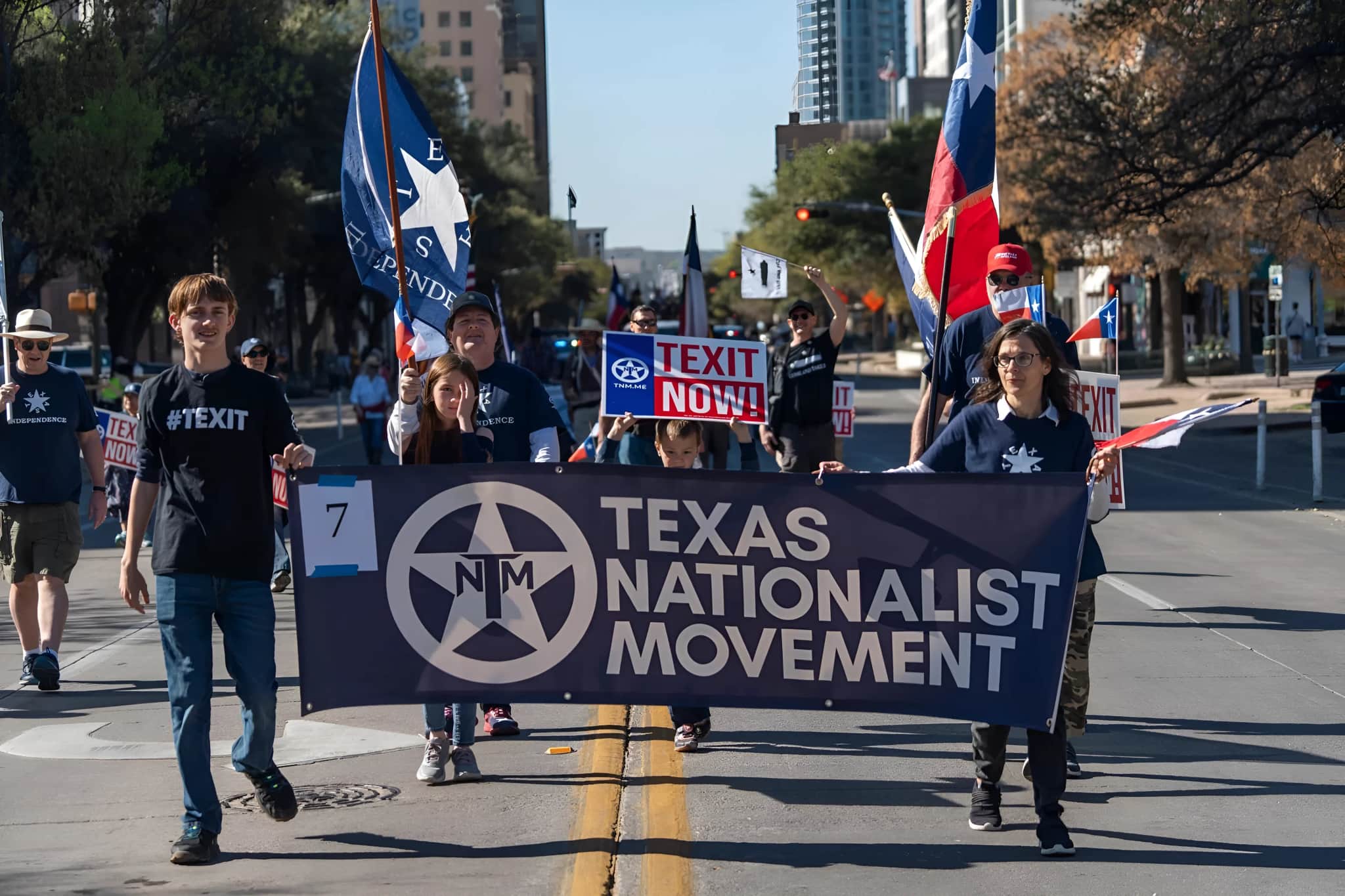 Texas Nationalist Movement members marching down a Texas street with a TNM banner and Texas independence flags.