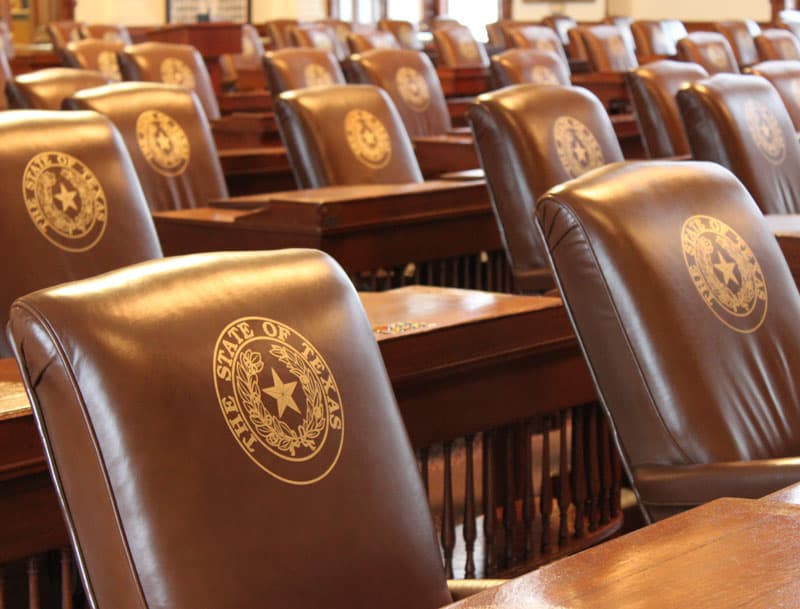 Empty leather chair-backs in the Texas House of Representatives chamber, each embossed with the gold State of Texas seal.